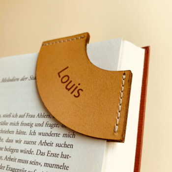 Lesezeichen aus hellem Natur Leder mit einer Gravur "Louis". Das Lesezeichen ist auf eine Buchseite gesteckt.